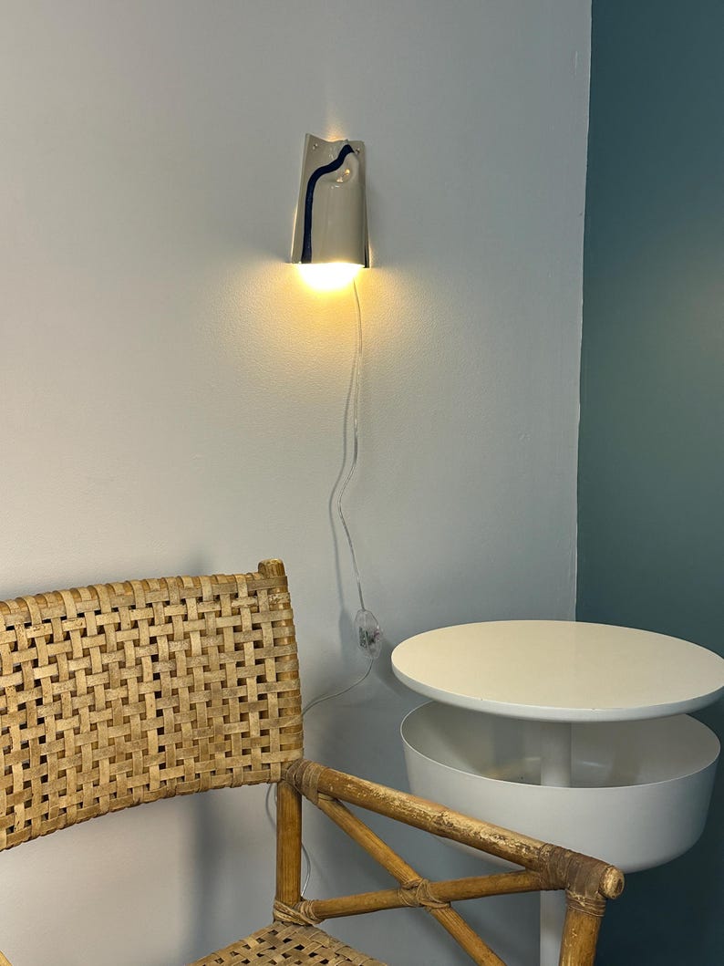 May include: A wall-mounted light fixture with a conical, off-white ceramic shade featuring a single vertical dark blue painted stripe. The lamp emits a warm, downward-facing glow and includes a thin, transparent power cord with a small switch.