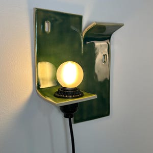 May include: A wall-mounted sconce featuring a glossy, dark green ceramic backplate with an irregular, folded shape. A black socket holds a small, glowing, frosted spherical light bulb. A black power cord extends downward from the base of the socket.