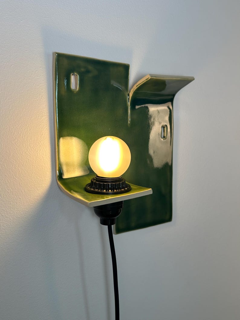 May include: A wall-mounted sconce featuring a glossy, dark green ceramic backplate with an irregular, folded shape. A black socket holds a small, glowing, frosted spherical light bulb. A black power cord extends downward from the base of the socket.