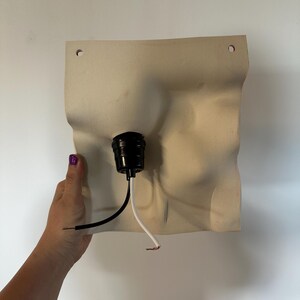 May include: A square, off-white wall sconce base with a wavy, organic surface texture. It features two small circular holes at the top for mounting and a central black light socket fixture with one black and one white electrical wire extending out.