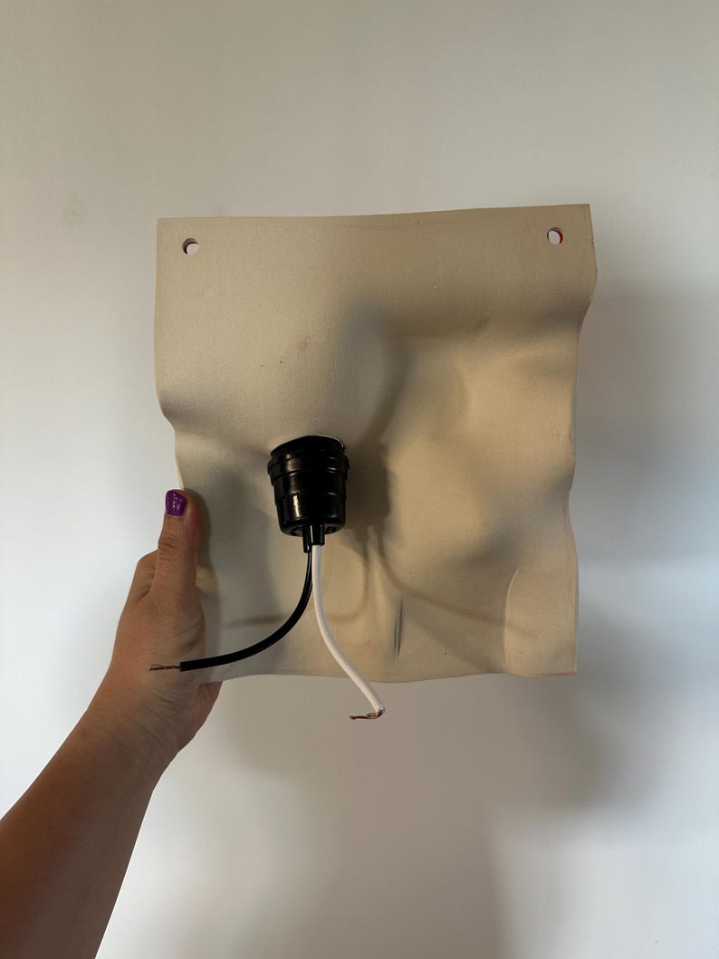 May include: A square, off-white wall sconce base with a wavy, organic surface texture. It features two small circular holes at the top for mounting and a central black light socket fixture with one black and one white electrical wire extending out.