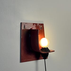 May include: A wall-mounted sconce featuring a glossy, dark reddish-brown ceramic backplate with a curved, protruding shelf. A black socket holds a round, frosted white light bulb that emits a warm glow, connected by a black power cord.