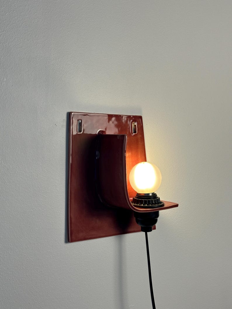 May include: A wall-mounted sconce featuring a glossy, dark reddish-brown ceramic backplate with a curved, protruding shelf. A black socket holds a round, frosted white light bulb that emits a warm glow, connected by a black power cord.