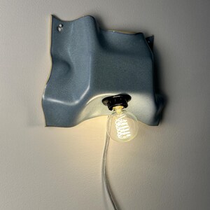 May include: A wall-mounted light fixture featuring a wavy, blue ceramic shade with a matte finish. A black socket holds a clear, round Edison-style light bulb with a visible glowing filament. A thin, transparent power cord hangs from the bottom.