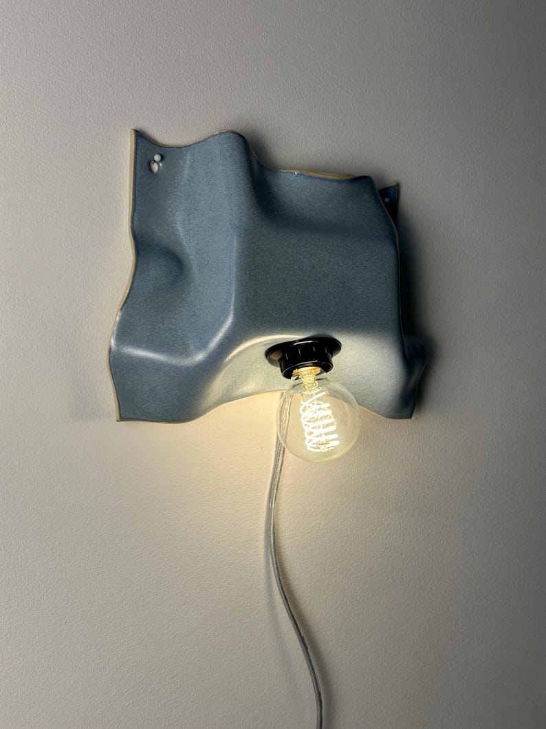 May include: A wall-mounted light fixture featuring a wavy, blue ceramic shade with a matte finish. A black socket holds a clear, round Edison-style light bulb with a visible glowing filament. A thin, transparent power cord hangs from the bottom.