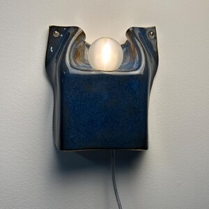 May include: A wall-mounted ceramic sconce featuring a deep blue, glossy glaze with an organic, curved shape. A single, spherical, frosted glass light bulb sits in the center indentation, emitting a soft glow. A thin power cord extends downward.