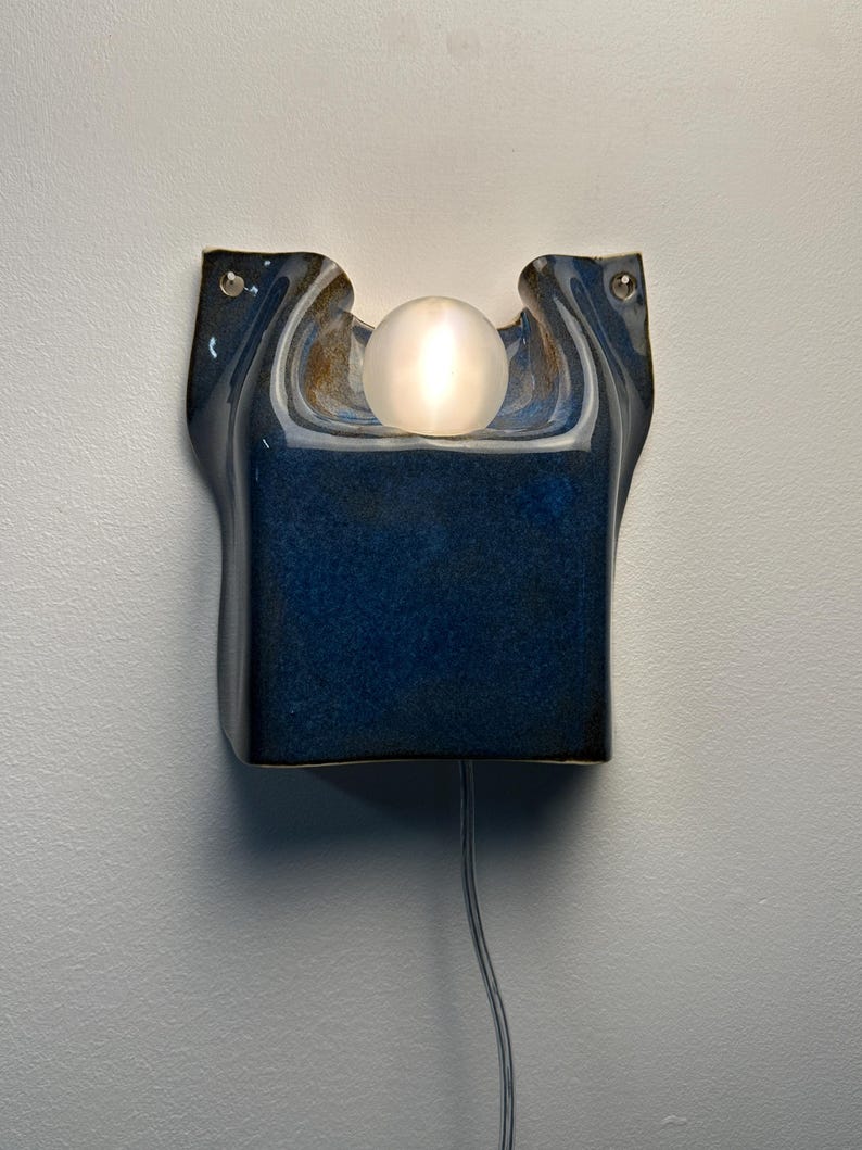 May include: A wall-mounted ceramic sconce featuring a deep blue, glossy glaze with an organic, curved shape. A single, spherical, frosted glass light bulb sits in the center indentation, emitting a soft glow. A thin power cord extends downward.