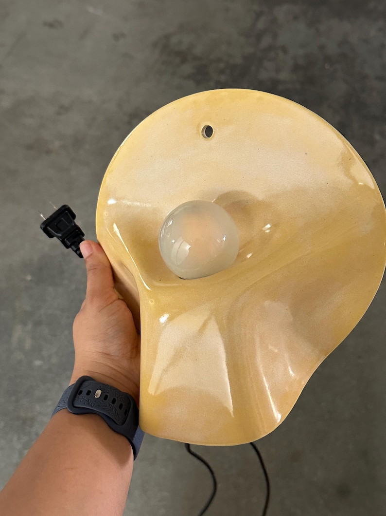 May include: A wall-mounted lamp featuring a glossy, pale yellow, shell-shaped ceramic shade with a smooth, undulating surface. A single, round, frosted glass light bulb is centered within the shade, and a black electrical cord with a plug is attached.