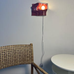 May include: A wall-mounted sconce featuring a glossy, deep burgundy ceramic shade with an undulating, wave-like form. The fixture emits a warm, glowing light from a single exposed bulb, connected by a thin, light-colored electrical cord.