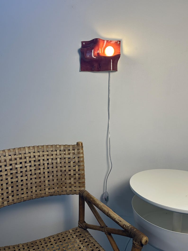 May include: A wall-mounted sconce featuring a glossy, deep burgundy ceramic shade with an undulating, wave-like form. The fixture emits a warm, glowing light from a single exposed bulb, connected by a thin, light-colored electrical cord.