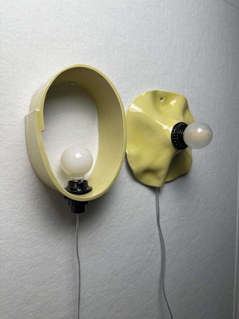 May include: Two pale yellow wall-mounted light fixtures. One features an oval-shaped frame with a central white bulb, while the other has a ruffled, organic-shaped ceramic shade with a matching white bulb. Both include black sockets and thin cords.