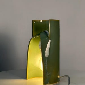 May include: A table lamp with a glossy, olive-green ceramic body featuring a curved, sculptural design. The lamp has an open front section that reveals a single glowing light bulb inside, and a thin, light-colored electrical cord extends from the base.