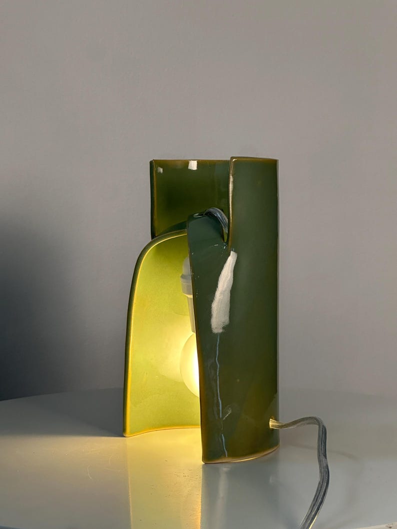 May include: A table lamp with a glossy, olive-green ceramic body featuring a curved, sculptural design. The lamp has an open front section that reveals a single glowing light bulb inside, and a thin, light-colored electrical cord extends from the base.