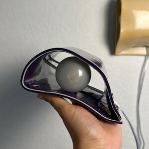 May include: A curved, deep purple ceramic wall sconce featuring a glossy finish and a central circular opening. A round, frosted white light bulb is positioned within the hollow center of the fixture, which includes a clear electrical cord.