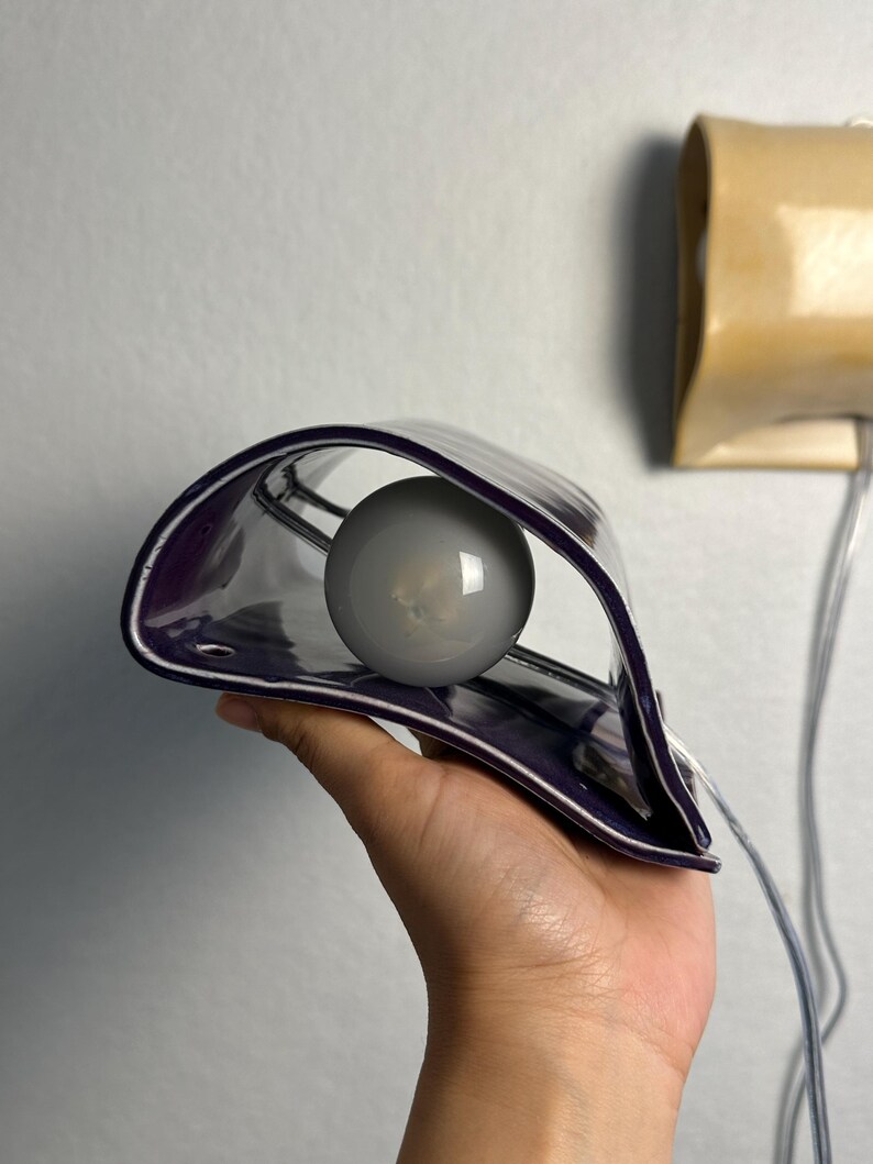 May include: A curved, deep purple ceramic wall sconce featuring a glossy finish and a central circular opening. A round, frosted white light bulb is positioned within the hollow center of the fixture, which includes a clear electrical cord.
