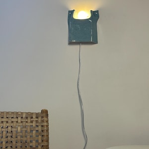 May include: A wall-mounted ceramic sconce with a speckled light blue glaze and a U-shaped top opening. The fixture emits a warm, glowing light from a single bulb housed within the ceramic body and includes a long, thin, light-colored power cord.