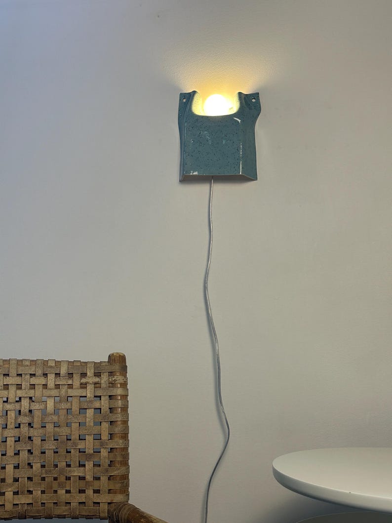 May include: A wall-mounted ceramic sconce with a speckled light blue glaze and a U-shaped top opening. The fixture emits a warm, glowing light from a single bulb housed within the ceramic body and includes a long, thin, light-colored power cord.
