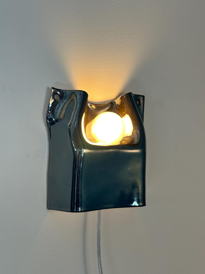May include: A wall-mounted lamp featuring a glossy, dark metallic ceramic housing with an irregular, fluid shape. The fixture includes a central circular opening that emits a warm, glowing light from an internal bulb, with mounting holes on the sides.