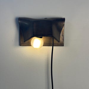 Pode incluir: Um candeeiro de parede com um abajur retangular de metal em acabamento mate escuro e bordas curvas em forma de asa. Uma l&acirc;mpada redonda fosca emite uma luz quente por baixo do abajur, com um cabo de alimenta&ccedil;&atilde;o preto pendurado verticalmente.