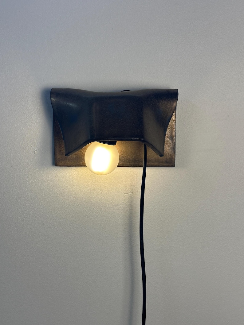 Pode incluir: Um candeeiro de parede com um abajur retangular de metal em acabamento mate escuro e bordas curvas em forma de asa. Uma l&acirc;mpada redonda fosca emite uma luz quente por baixo do abajur, com um cabo de alimenta&ccedil;&atilde;o preto pendurado verticalmente.