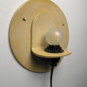 Pu&ograve; includere: Un'applique circolare color giallo pallido con una piastra posteriore rotonda e piatta e un piccolo ripiano sporgente. Un portalampada nero &egrave; montato sul ripiano e sostiene una piccola lampadina sferica smerigliata. Un cavo nero pende verso il basso.