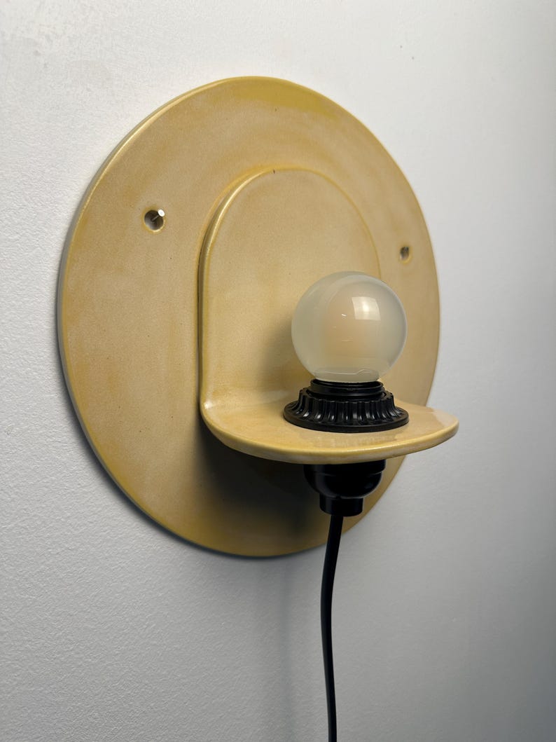 Pu&ograve; includere: Un'applique circolare color giallo pallido con una piastra posteriore rotonda e piatta e un piccolo ripiano sporgente. Un portalampada nero &egrave; montato sul ripiano e sostiene una piccola lampadina sferica smerigliata. Un cavo nero pende verso il basso.