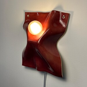 Puede incluir: Una l&aacute;mpara de pared con una forma de cer&aacute;mica roja profunda, brillante y ondulada, con una superficie lisa y sinuosa. Una bombilla blanca esf&eacute;rica y brillante est&aacute; colocada en una curva c&oacute;ncava de la estructura de cer&aacute;mica roja.