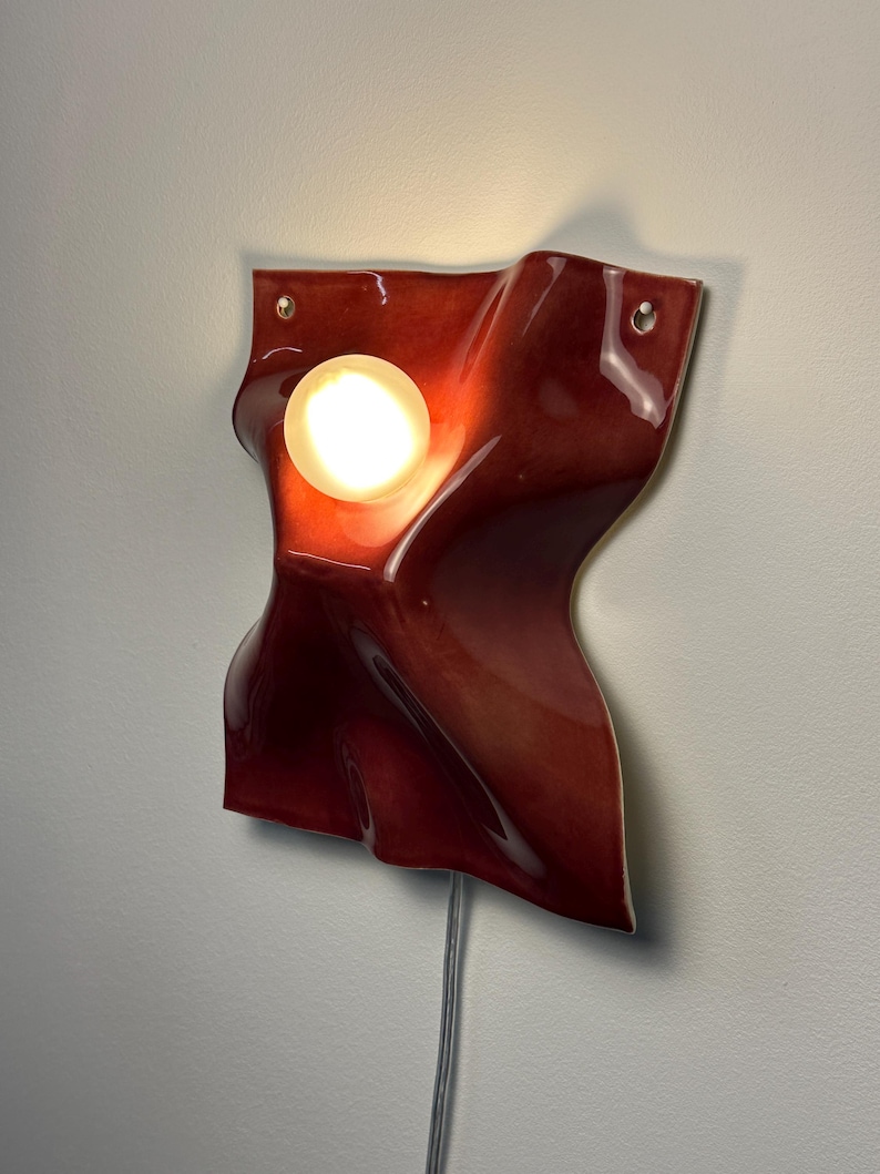 Puede incluir: Una l&aacute;mpara de pared con una forma de cer&aacute;mica roja profunda, brillante y ondulada, con una superficie lisa y sinuosa. Una bombilla blanca esf&eacute;rica y brillante est&aacute; colocada en una curva c&oacute;ncava de la estructura de cer&aacute;mica roja.