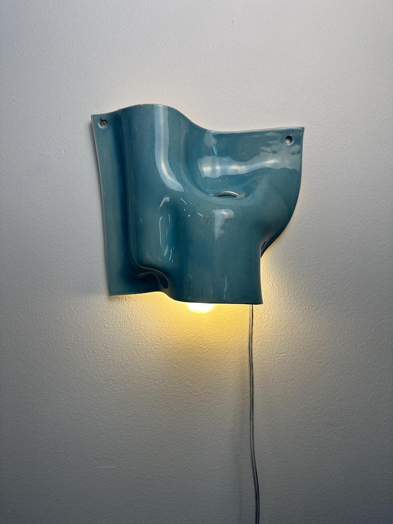 May include: A wall-mounted light fitting with a glossy, blue, undulating ceramic shade. The sculptural form features soft curves and two small circular holes for mounting. A warm, glowing light bulb is visible at the bottom, with a thin power cord.