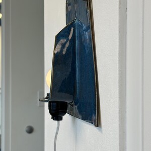 May include: A modern wall sconce with a dark blue, glossy ceramic shade and a brass-colored trim. The sconce features a black light socket and a clear cord, mounted on a white wall. The light bulb is visible and lit.