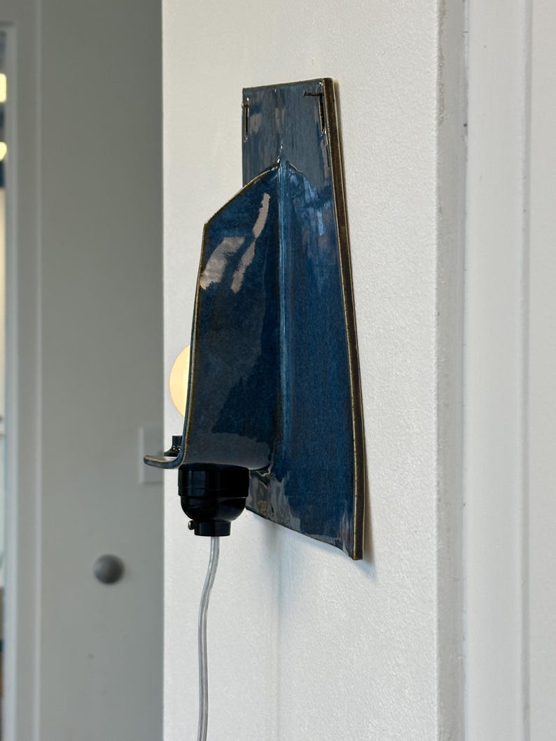 May include: A modern wall sconce with a dark blue, glossy ceramic shade and a brass-colored trim. The sconce features a black light socket and a clear cord, mounted on a white wall. The light bulb is visible and lit.