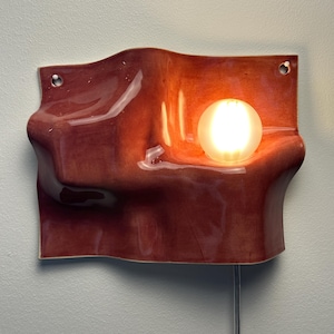May include: A wall-mounted ceramic light fixture with a glossy, wavy, terracotta-colored surface. The piece features two circular holes for mounting and a single, glowing, spherical light bulb nestled within a curved indentation on the right side.