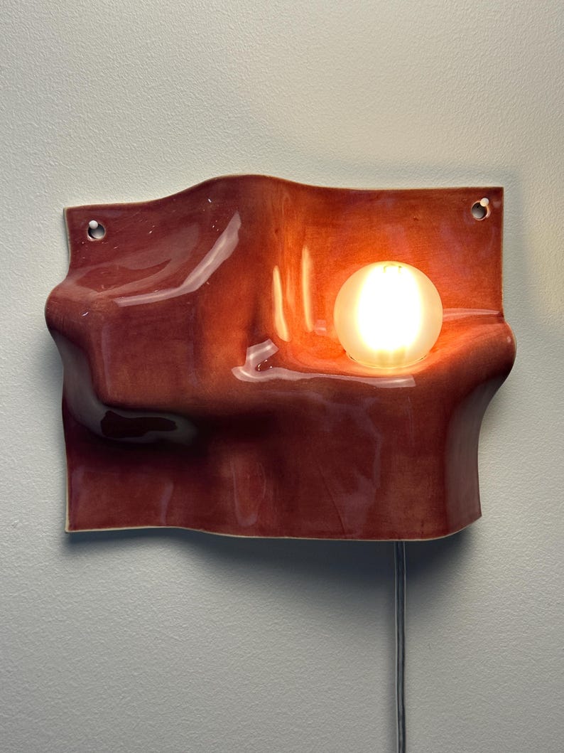 May include: A wall-mounted ceramic light fixture with a glossy, wavy, terracotta-colored surface. The piece features two circular holes for mounting and a single, glowing, spherical light bulb nestled within a curved indentation on the right side.