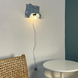 May include: A wall-mounted light fixture with a glossy, light blue, irregularly shaped ceramic shade. The fixture features a single exposed light bulb emitting a warm glow, connected to a long white power cord with an integrated toggle switch.