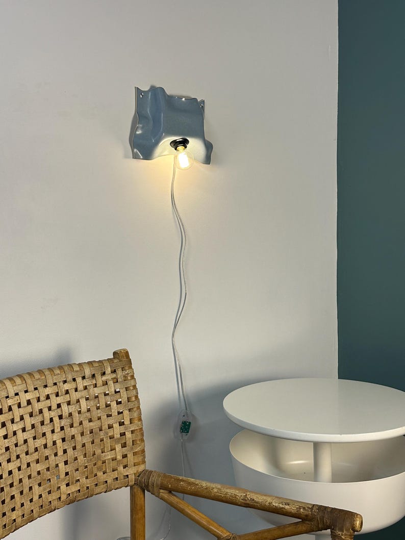 May include: A wall-mounted light fixture with a glossy, light blue, irregularly shaped ceramic shade. The fixture features a single exposed light bulb emitting a warm glow, connected to a long white power cord with an integrated toggle switch.