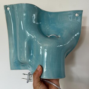 May include: A light blue ceramic wall lamp with a wavy, organic shape and a glossy finish. The fixture features two circular holes at the top corners for mounting and includes a clear electrical cord with a two-pin plug attached at the base.