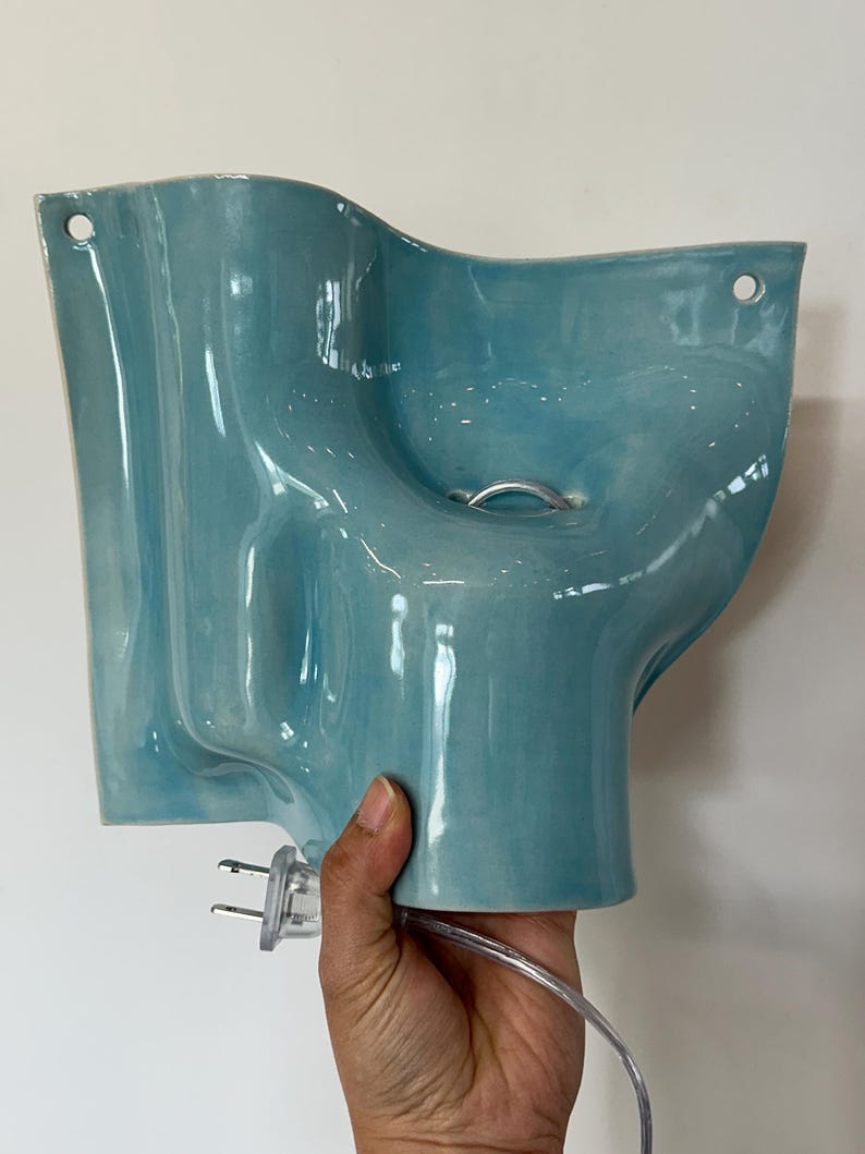 May include: A light blue ceramic wall lamp with a wavy, organic shape and a glossy finish. The fixture features two circular holes at the top corners for mounting and includes a clear electrical cord with a two-pin plug attached at the base.