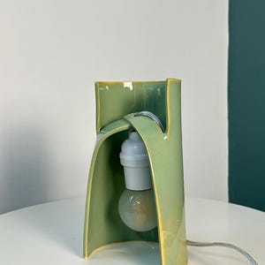 May include: A sage green ceramic table lamp with a curved, sculptural form. The lamp features a glossy finish with yellow-toned edges, a white light bulb socket, and a clear light bulb suspended within the open, arched structure of the ceramic body.