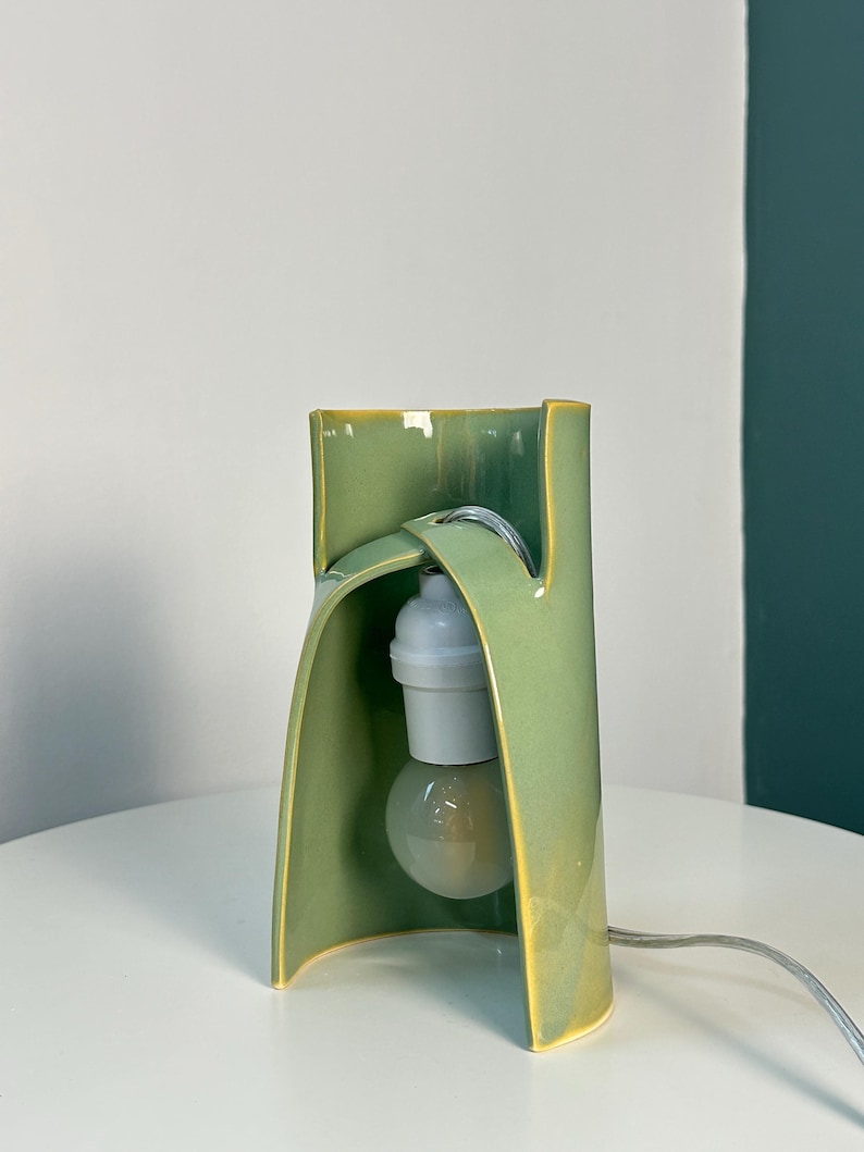 May include: A sage green ceramic table lamp with a curved, sculptural form. The lamp features a glossy finish with yellow-toned edges, a white light bulb socket, and a clear light bulb suspended within the open, arched structure of the ceramic body.