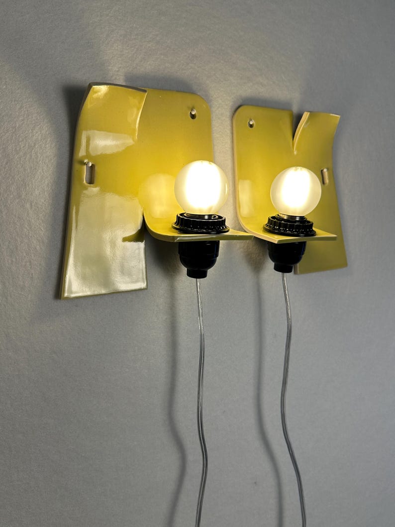 May include: Two wall-mounted light fixtures featuring glossy yellow ceramic backplates with irregular, organic shapes. Each fixture includes a black socket holding a round, frosted white light bulb, with a thin, clear electrical cord extending downward.
