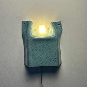 May include: A light blue wall sconce with a speckled finish and a central cutout holding a glowing, frosted spherical light bulb. The ceramic fixture features two mounting holes at the top corners and a thin black power cord extending downward.
