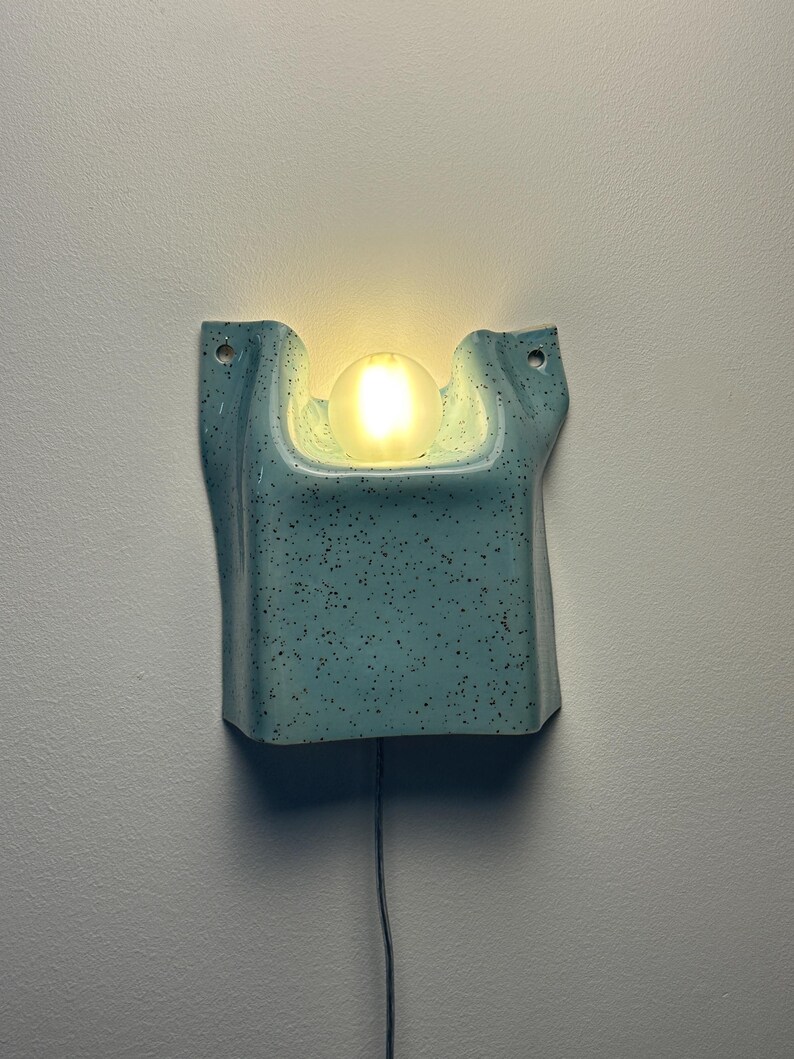 May include: A light blue wall sconce with a speckled finish and a central cutout holding a glowing, frosted spherical light bulb. The ceramic fixture features two mounting holes at the top corners and a thin black power cord extending downward.