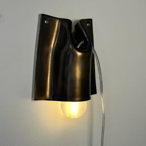 May include: A wall-mounted lamp featuring a dark, metallic, curved shade with a smooth finish. The fixture has two circular mounting holes at the top and a single, glowing, frosted spherical light bulb extending from the bottom. A clear power cord hangs down from the side of the lamp.
