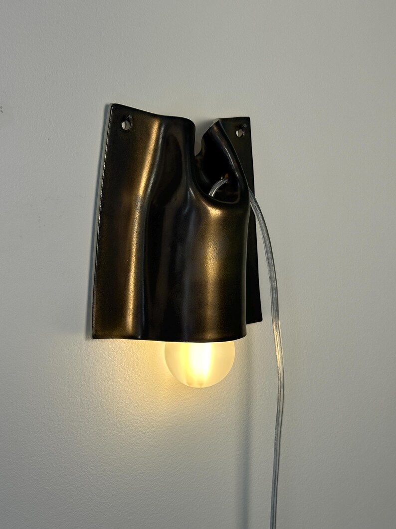 May include: A wall-mounted lamp featuring a dark, metallic, curved shade with a smooth finish. The fixture has two circular mounting holes at the top and a single, glowing, frosted spherical light bulb extending from the bottom. A clear power cord hangs down from the side of the lamp.