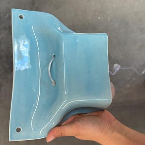 K&ouml;nnte beinhalten: Eine hellblaue Wandleuchte aus Keramik mit gl&auml;nzender Oberfl&auml;che. Das St&uuml;ck hat eine geschwungene, ausgestellte Form mit zwei runden Befestigungsl&ouml;chern oben und unten sowie einer zentralen &Ouml;ffnung f&uuml;r eine Lampenfassung mit transparentem Kabel.