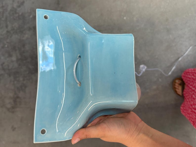 K&ouml;nnte beinhalten: Eine hellblaue Wandleuchte aus Keramik mit gl&auml;nzender Oberfl&auml;che. Das St&uuml;ck hat eine geschwungene, ausgestellte Form mit zwei runden Befestigungsl&ouml;chern oben und unten sowie einer zentralen &Ouml;ffnung f&uuml;r eine Lampenfassung mit transparentem Kabel.