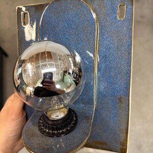 May include: A wall-mounted light fixture featuring a rectangular blue ceramic backplate with rounded edges and two rectangular cutouts. A circular ceramic base extends from the center, holding a black socket and a large, round, silver-tinted light bulb.