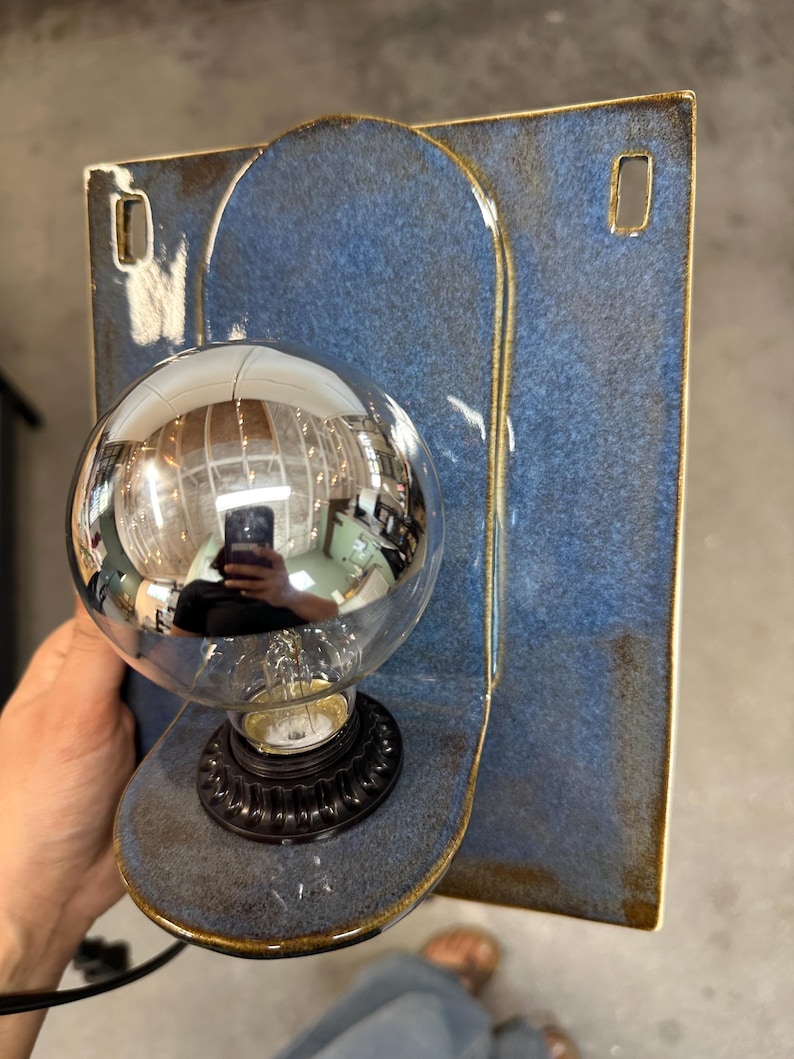 May include: A wall-mounted light fixture featuring a rectangular blue ceramic backplate with rounded edges and two rectangular cutouts. A circular ceramic base extends from the center, holding a black socket and a large, round, silver-tinted light bulb.