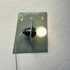 May include: A wall-mounted light fixture featuring a rectangular, speckled, light blue-green ceramic backplate with two small mounting holes. A black socket holds a glowing, round, frosted white light bulb, connected by a clear electrical cord.