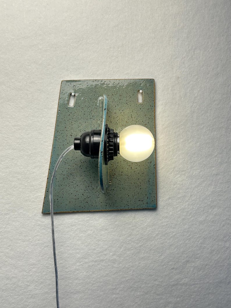 May include: A wall-mounted light fixture featuring a rectangular, speckled, light blue-green ceramic backplate with two small mounting holes. A black socket holds a glowing, round, frosted white light bulb, connected by a clear electrical cord.